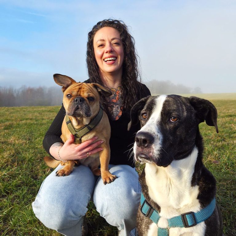 Hi, mein Name ist... Frau mit lockigem Haar und Tattoo im Dekolletee hockt im Gras und hält einen Hund, neben ihr ein weiterer Hund.